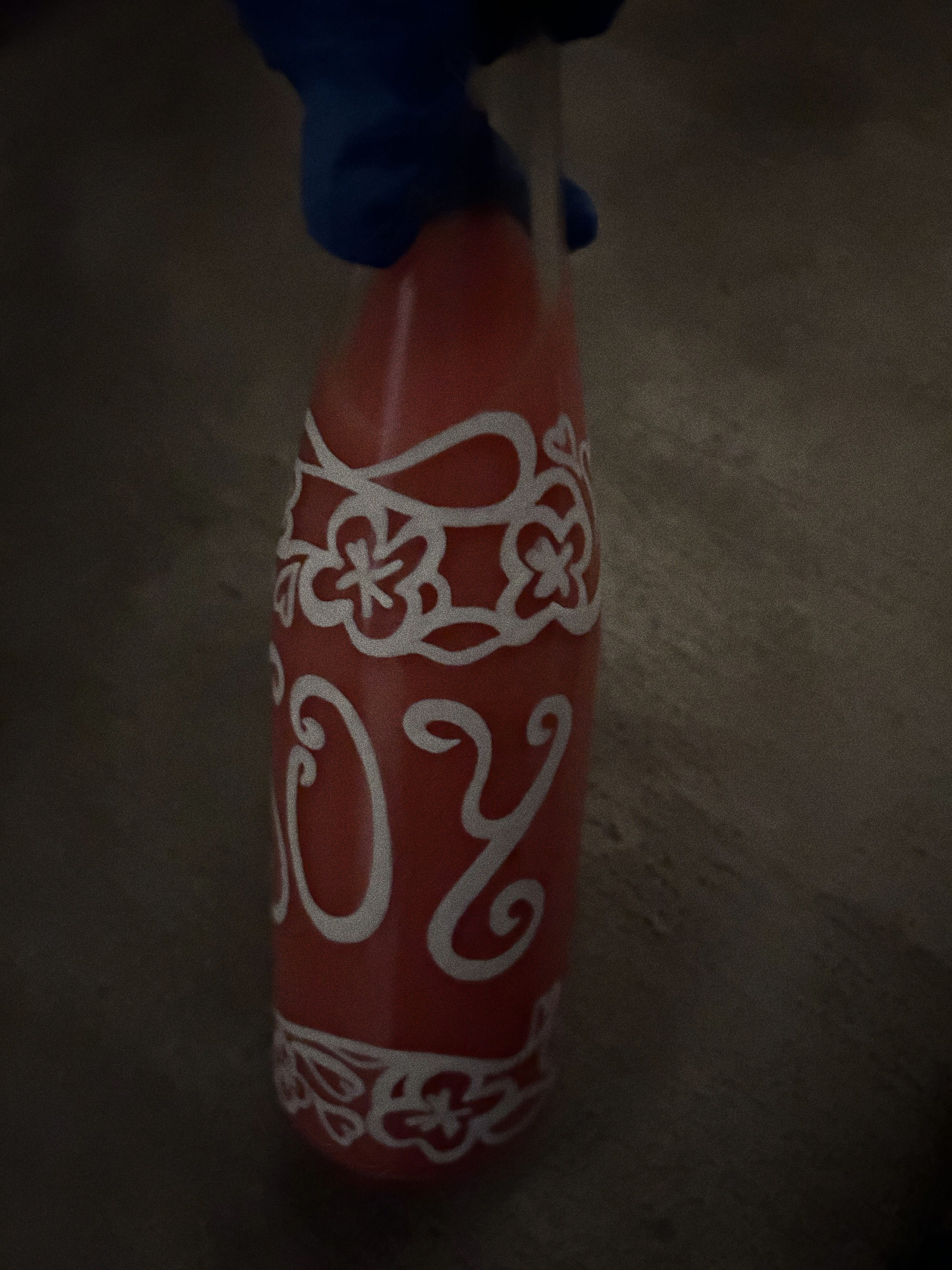 A blue latex gloved hand holding a glass bottle of 'Joy'. On the bottle is white flower detailing alongside the title, and inside the bottle is a pink liquid. Behind the bottle is a concrete floor.